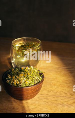 Chamomile tea and dry chamomile flowers in jar. Medicine camomile in sunlight. Eco products. Glass of natural tea and herbs. Herbal drinks. Stock Photo