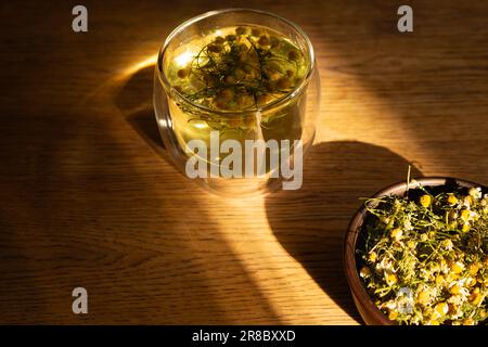 Chamomile tea and dry chamomile flowers in jar. Medicine camomile in sunlight. Eco products. Glass of natural tea and herbs. Herbal drinks. Stock Photo