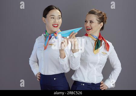 smiling elegant female air hostesses in blue skirt, white shirt and scarf with paper airplane against gray background. Stock Photo