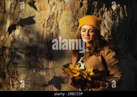 Hello november. pensive elegant female in beige coat and orange hat with autumn yellow leaves against huge tree outside on the city park in autumn. Stock Photo