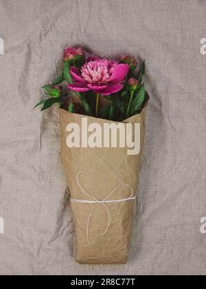 Pink peony flowers bouquet in kraft paper on grey linen fabric background top view, Minimal flat lay holidays celebrating concept.Women's Mother's Day Stock Photo
