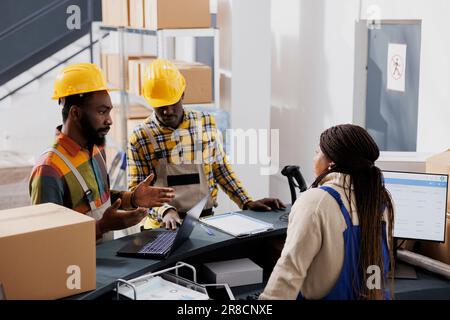 Warehouse workers asking postal manager about merchandise supply schedule. Industrial storehouse employees using laptop and talking with supervisor at reception counter desk Stock Photo