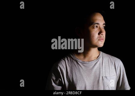 Low key shot of a young Asian man wearing a grey T-shirt and a silver necklace is looking at the right side of the camera. Isolated black background. Stock Photo