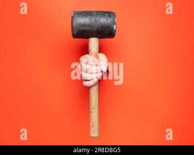 A hand is holding a big rubber mallet through torn orange paper. No face, copy space, concept. Stock Photo