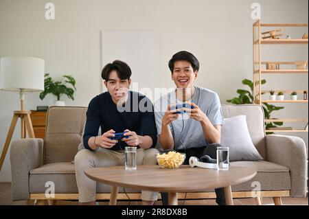 Two overjoyed and happy young Asian male friends are playing video games console on a sofa in the living room together. joysticks, video game controll Stock Photo