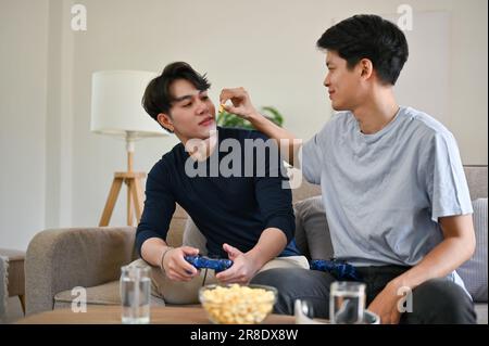A happy and joyful young Asian gay man feeds popcorn to his boyfriend while having fun in the living room together. LGBTQ+, gay couple, lifestyle Stock Photo