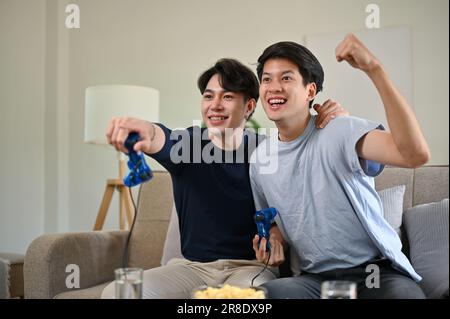 Two overjoyed young Asian male friends raised their hands, held game controllers, looked at the TV screen, and celebrated their game wins while playin Stock Photo
