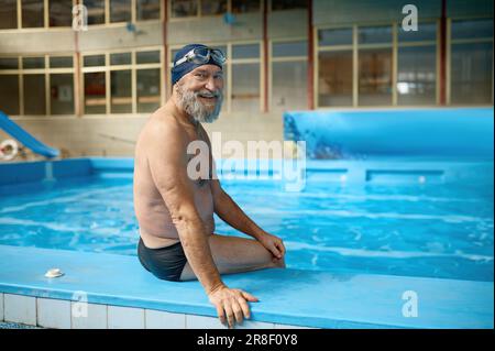 Relaxed senior man in swimming hat and goggles on head sitting at poolside Stock Photo