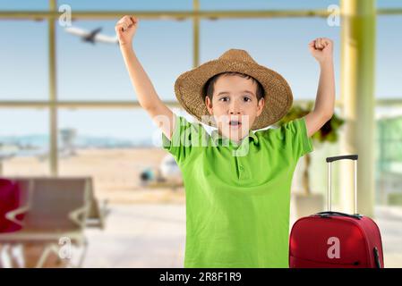 Child on vacation wearing green t-shirt summer hat at airport, very happy and excited doing winner gesture with arms raised, smiling and screaming for Stock Photo