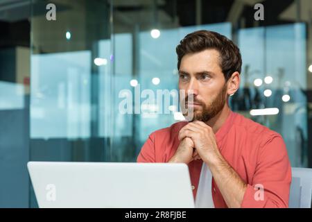 Portrait of a young thoughtful serious man businessman, freelancer, programmer sitting at a table, working on a laptop, folded his hands together, looks to the side. Stock Photo