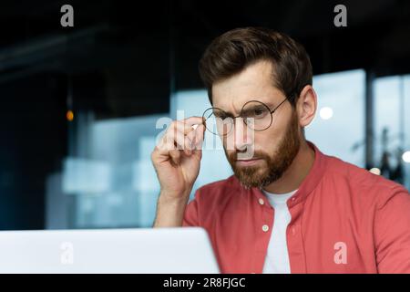Close-up photo. Portrait of a young male programmer, a businessman who is concentrating on working in the office at a laptop, looking at the monitor, holding glasses on his face. Stock Photo