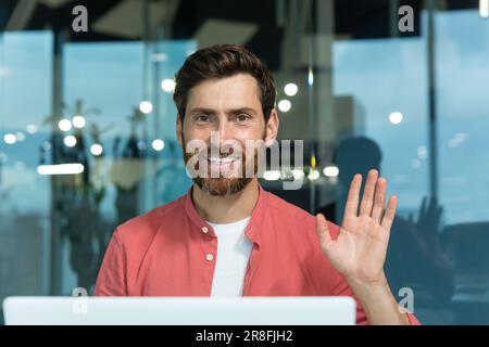Portrait of a young male programmer in a red shirt working in the office at a laptop. He looks at the camera with a smile, waves his hand. Close-up photo. Stock Photo
