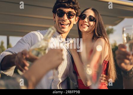 A young Caucasian couple, dressed in casual-elegant attire, embracing and smiling at an outdoor party. The man is pouring champagne for friends. Stock Photo