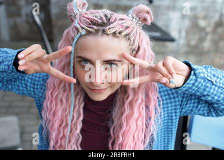 An attractive girl with pink hair and a blue shirt drinks coffee and listens to music sitting at a table in a cafe. A girl with long pink hair is havi Stock Photo