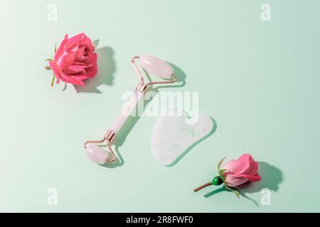 Face roller, gua sha stone and pink rose flowers on light green background with hard shadows. Skin care, beauty treatment concept. Top view, flat lay. Stock Photo