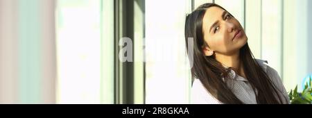 Dreamy young business woman is sitting on windowsill near huge window in modern office sun is shining brightly Stock Photo
