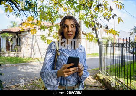 young latina woman of argentinian ethnicity, standing outside her home on the sidewalk with her phone in her hand, thinking about the direction to ord Stock Photo