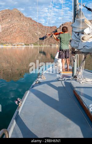 Early in the morning a young man stands on the bow of his sailboat playing the violin, Sea of Cortez, Bahia Concepcion, Baja California Sur, Mexico. Stock Photo