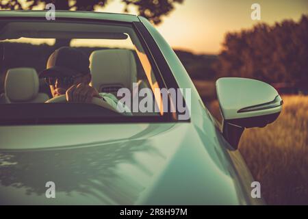 Front View of Wealthy Caucasian Man in Baseball Cap and Sunglasses Driving Modern White Convertible at Sunset. Stock Photo