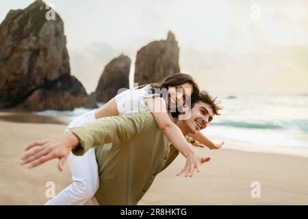 Happy loving young couple enjoying date at beach, husband giving his joyful wife a piggyback, walking by the seashore Stock Photo