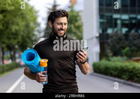 Portrait of mature successful adult athlete, bearded man going to workout, coach holding fitness mat and phone, browsing online app for joint physical training. Stock Photo