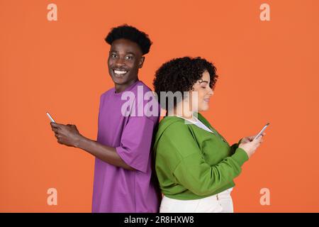 Cheerful interracial young couple standing back to back, using phones Stock Photo