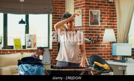 Modern young adult ironing clothes and getting angry at life partner, needing help with household chores. Stressed exhausted girlfriend cleaning apartment and doing housekeeping without man. Stock Photo