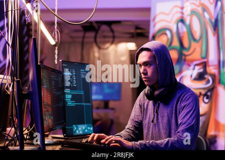 Asian hacker cracking password to get illegal access to database confidential information. Young man in hood infecting victim computer with internet virus in abandoned warehouse Stock Photo