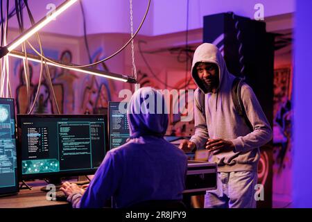 African american hacker bringing black box with technology for cyberattack. Young man in hood showing case with tool for data breach and explaining internet crime plan in hideout place Stock Photo