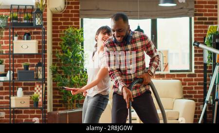 Young life partners vacuuming floors and showing dance moves at home, having fun doing housework. Cheerful modern man and woman dancing and singing on music, acting silly. Handheld shot. Stock Photo