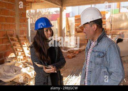 Male engineer is talking with female architect, during work on construction site, both wearing safety helmet, and looking at blueprint on tablet at co Stock Photo