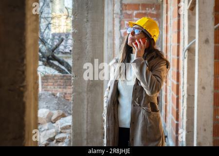 Portrait of successful female builder with yellow safety helmet, making a call with her cell phone. Portrait of female architect standing looking at c Stock Photo