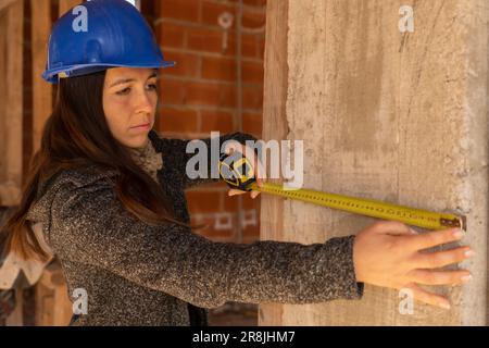 Concentrated handywoman with blue hard hat and tape measure measuring a wall on construction site. Copy space. Horizontal portrait of female architect Stock Photo