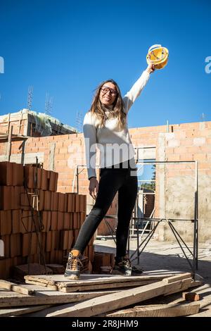 Successful mature female engineer. Vertical portrait of female architect standing smiling and looking at camera with her yellow hard hat holding it up Stock Photo