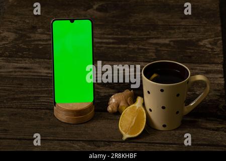 mobile phone with a green screen and a cup of tea with lemon on an wooden table Stock Photo
