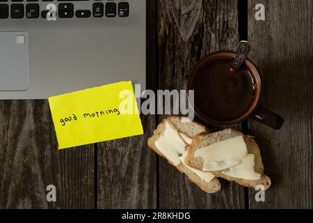 coffee and white bread and butter on an wooden table and the words in English good morning on yellow paper and a laptop Stock Photo