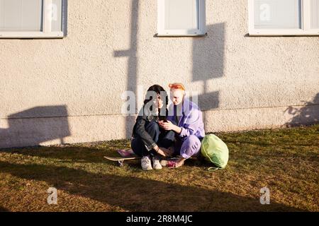 Smiling female friends or gay couple relaxing together while looking at cell phone Stock Photo