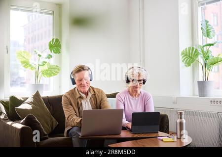 Senior man and woman sitting in living room and using laptop and digital tablet to edit podcast Stock Photo