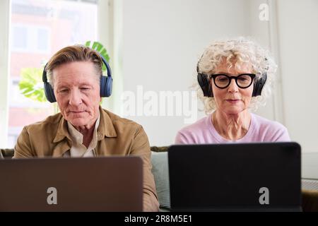 Senior man and woman sitting in living room and using laptop and digital tablet to edit podcast Stock Photo