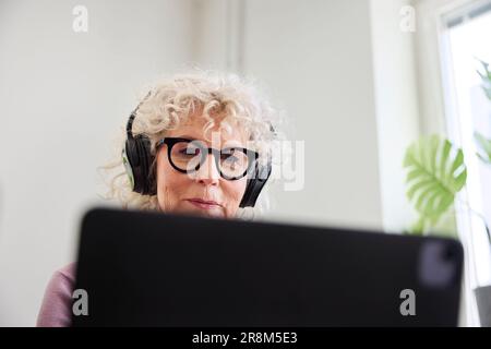 Smiling senior woman sitting withe headphones on and using laptop Stock Photo
