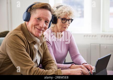 Smiling senior man looking at camera while wearing headphones, senior woman in background Stock Photo