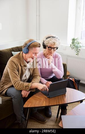 Senior man and woman sitting in living room and using laptop and digital tablet to edit podcast Stock Photo