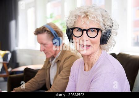 Smiling senior woman looking at camera while wearing headphones, senior man in background Stock Photo