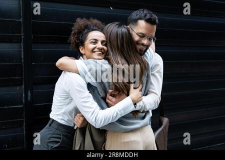 Cheerful businessman and businesswomen hugging each other in front of office building. Colleagues hug colleague who change her job and work, going to Stock Photo