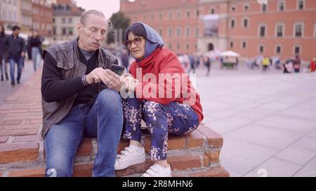 Elderly couple of tourists looking at their mobile phone are sitting in the historic center of an old European city and talking to each other. Palace Stock Photo