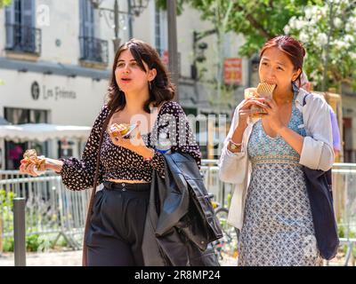 Two young casually dressed women eating snacks whilst walking through city center - Tours, Indre-et-Loire (37), France. Stock Photo