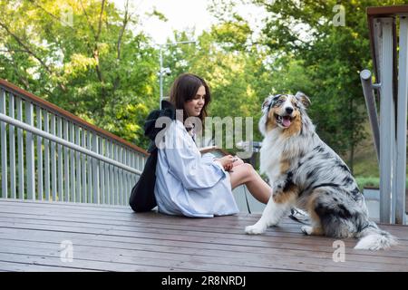 Young woman sitting next to her cute aussie dog in a park. Blue merle australian shepherd dog in urban park area next to female owner, walking pets in Stock Photo