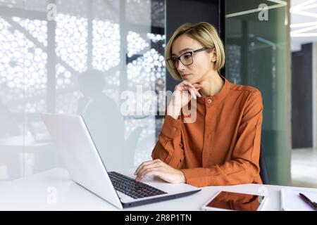 Serious thinking woman working with laptop inside office at workplace, businesswoman solving technical task, female programmer coding new software. Stock Photo