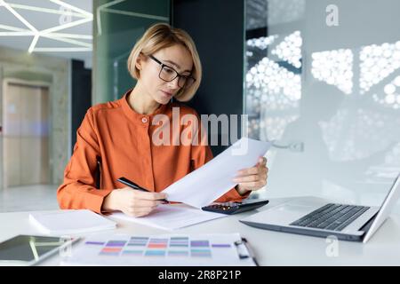 Serious and pensive business woman behind paper work inside office, female financier worker thinks about contracts and reports with charts and graphs, blonde successful woman uses laptop at work. Stock Photo