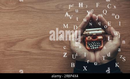 A hand holding toy typewriter with alphabet flying around on wooden table with copy space. Stock Photo